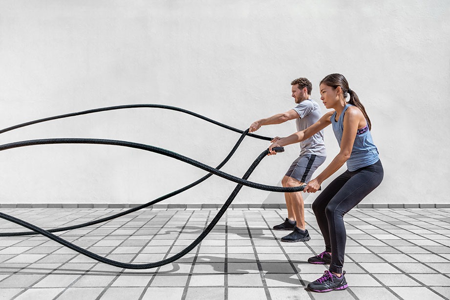 Fitness people exercising with battle ropes at gym. Woman and man couple training together doing battling rope workout working out arms and cardio for crossfit exercises.