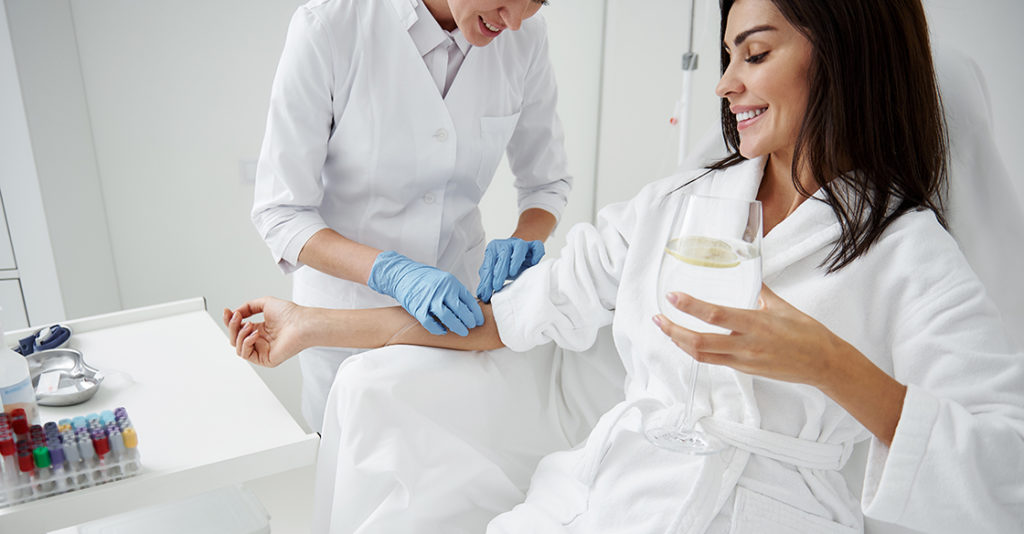 Cropped portrait of beautiful woman in white bathrobe sitting in armchair and receiving IV infusion. She is holding glass of beverage with lemon and smiling